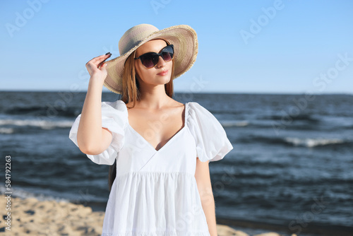 Happy blonde woman is posing on the ocean beach with sunglasses and a hat. Evening sun