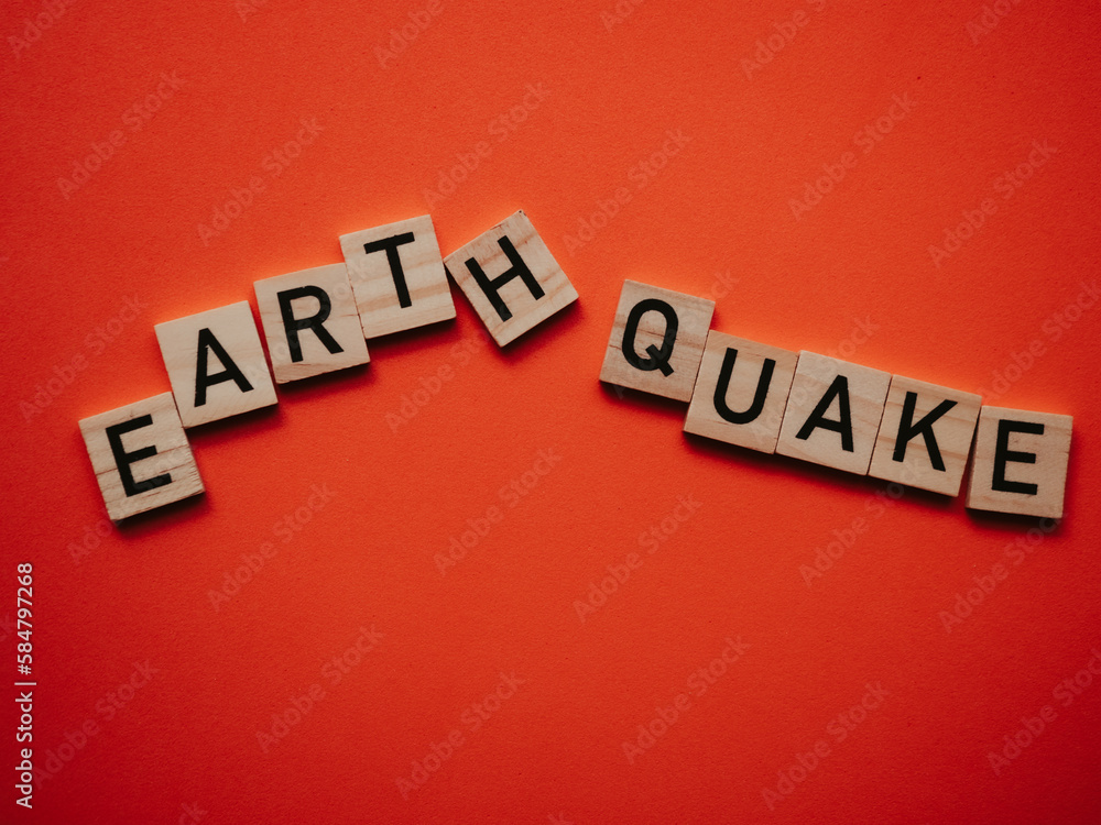 Obraz premium Inscription earthquake from vibration moves on background. Concept of earthquake, destruction and shifts of earth's crust. Wooden square blocks with letters are laid out on orange sheet. Slow motion