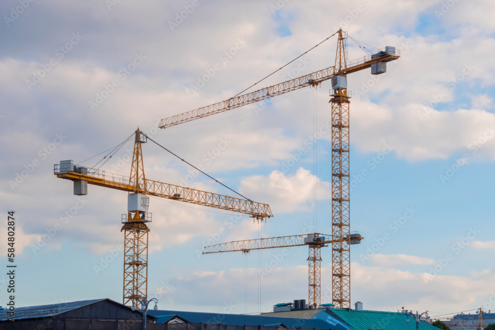 Three yellow tower construction cranes against summer cloudy blue sky with white clouds. Building process, architecture, urban, engineering, industrial and development concept