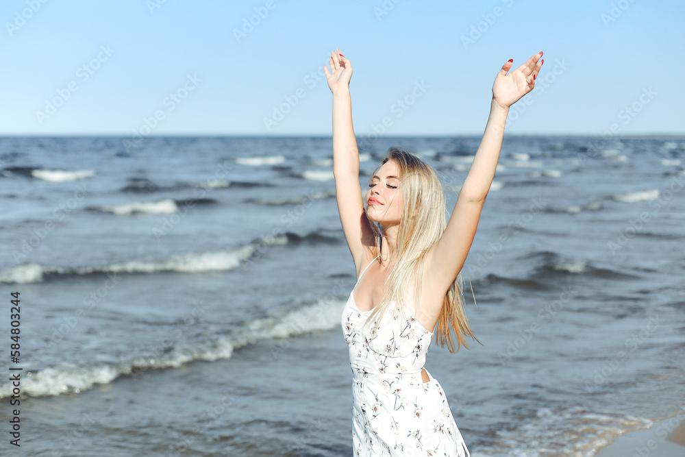 Happy blonde beautiful woman on the ocean beach standing in a white summer dress, raising hands Happy blonde beautiful woman on the ocean beach standing in a white summer dress, raising hands