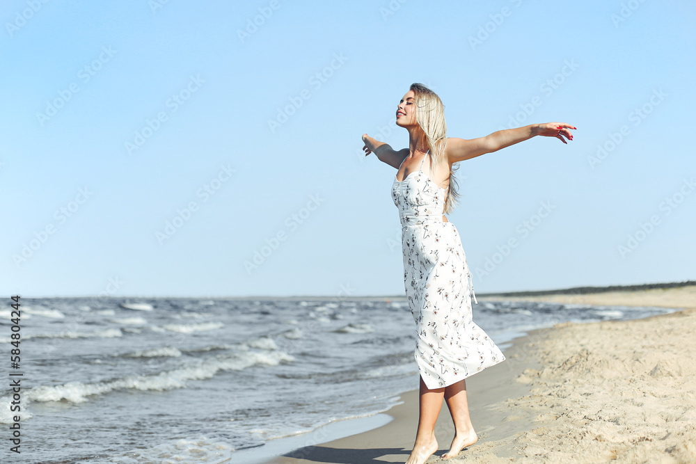 Happy blonde beautiful woman on the ocean beach standing in a white summer dress, open arms Happy blonde beautiful woman on the ocean beach standing in a white summer dress, open arms