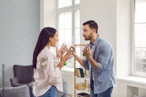 Angry young couple standing at home and arguing in the living room shouting on each other. Frustrated unhappy husband and wife quarreling. Bad marriage,relationships, quarrel and divorce concept.