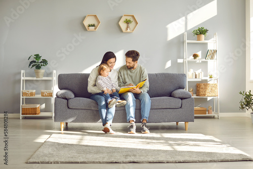 Smiling , happy mother and father with charming child engaged in educational activities. Loving family with their young child read a book together, sitting at home on cozy sofa. © Studio Romantic