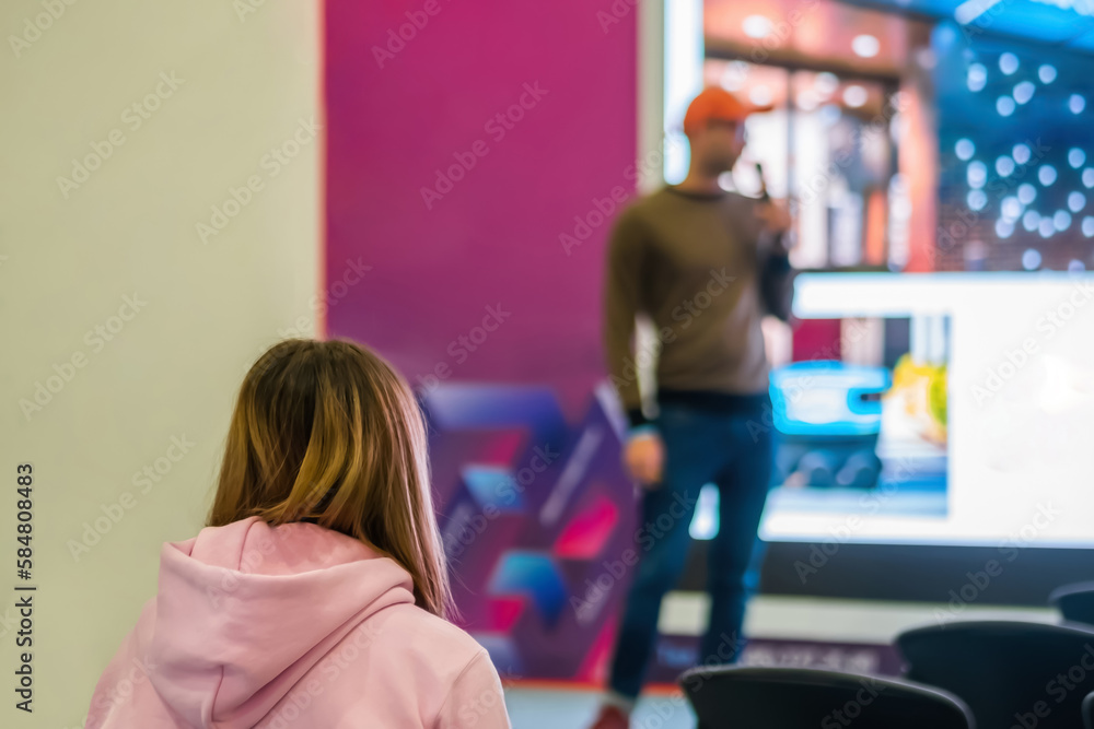 Back view of unrecognizable woman in pink hoody listening lecture, presentation at trade show, conference hall. Business, congress, seminar, corporate, training, education concept
