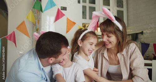 Joyful family of four painting Easter eggs together in bright modern kitchen. Parents and children wearing rabbit ears happily embrace and laugh. Family gathering in preparation for Easter.