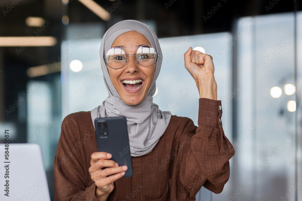 Closeup portrait of businesswoman in hijab, muslim celebrating victory and success good achievement results, workplace inside office looking at camera and happy, holding phone, received notification. - obrazy, fototapety, plakaty 
