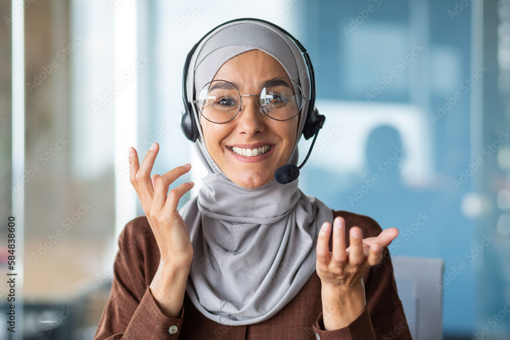 Online customer support worker consults a client remotely, a woman in a hijab with a headset for a video call talks, looks at the web camera and smiles at the interlocutor, closeup - obrazy, fototapety, plakaty 