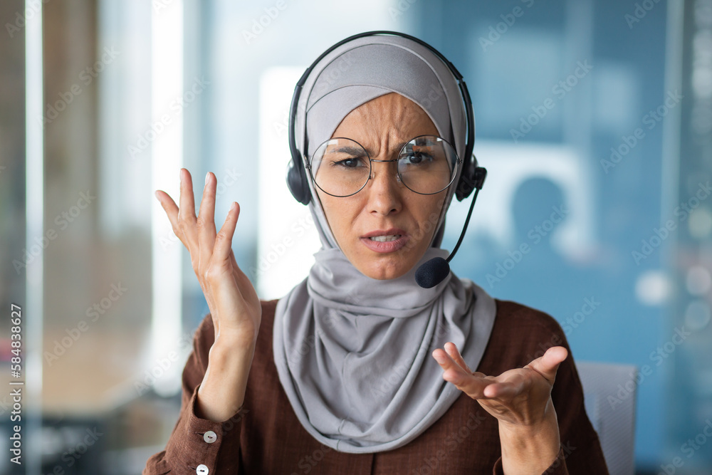 Angry woman in hijab talking on video call, businesswoman with headset phone close-up looking at web camera and talking to colleagues in online meeting. - obrazy, fototapety, plakaty 