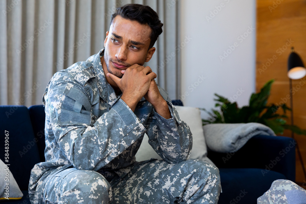 Fototapeta premium Thoughtful biracial male soldier wearing military uniform, sitting on sofa in living room at home