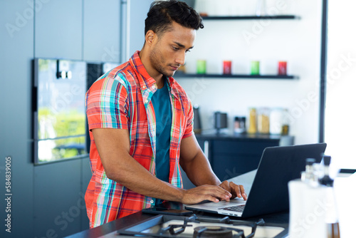 Concentrating biracial man leaning on countertop using laptop in kitchen
