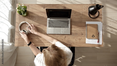 Top down view, girl in white t-shirt sits at laptop at home and works. young blonde woman puts on headphones and studies online courses in morning. Remote work of freelancer at home.