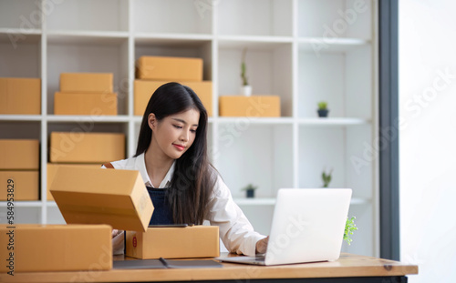 A portrait of a young Asian woman, e-commerce employee sitting in the office full of packages in the background write note of orders and a calculator, for SME business ecommerce and delivery business