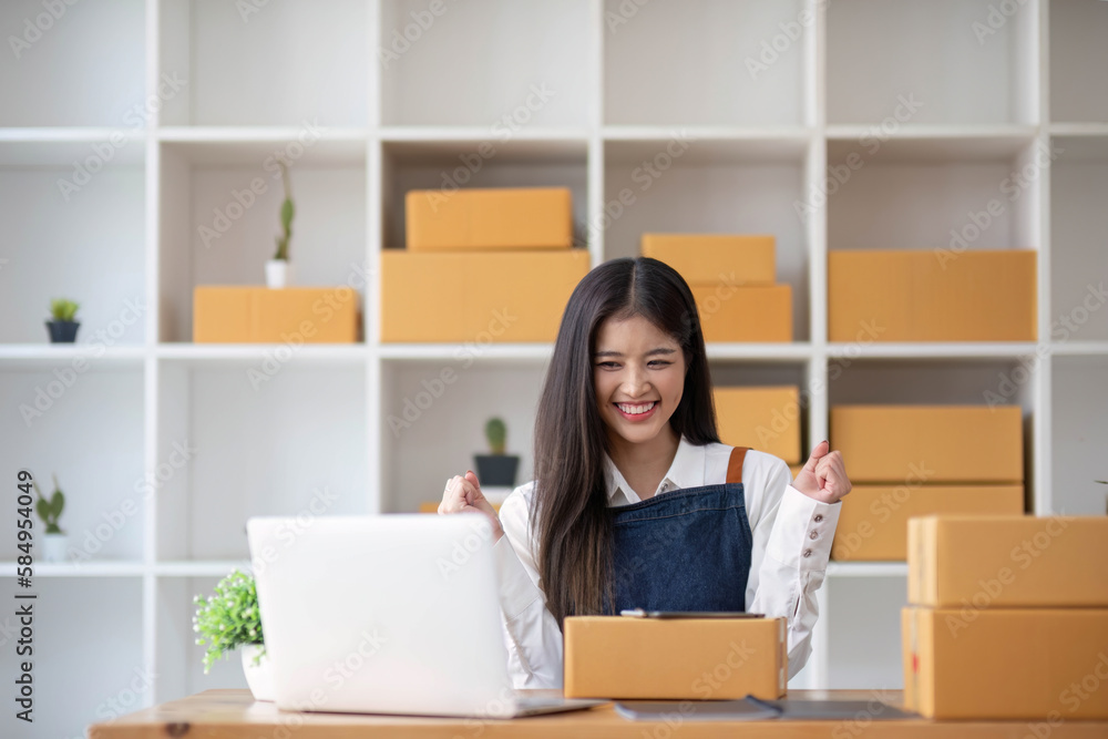 A portrait of a young Asian woman, e-commerce employee sitting in the office full of packages in the background write note of orders and a calculator, for SME business ecommerce and delivery business A portrait of a young Asian woman, e-commerce employee sitting in the office full of packages in the background write note of orders and a calculator, for SME business ecommerce and delivery business
