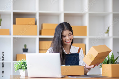 A portrait of a young Asian woman, e-commerce employee sitting in the office full of packages in the background write note of orders and a calculator, for SME business ecommerce and delivery business © wichayada