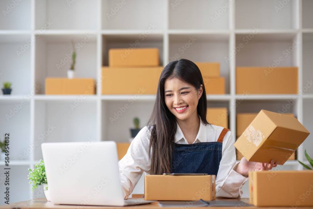 A portrait of a young Asian woman, e-commerce employee sitting in the office full of packages in the background write note of orders and a calculator, for SME business ecommerce and delivery business A portrait of a young Asian woman, e-commerce employee sitting in the office full of packages in the background write note of orders and a calculator, for SME business ecommerce and delivery business