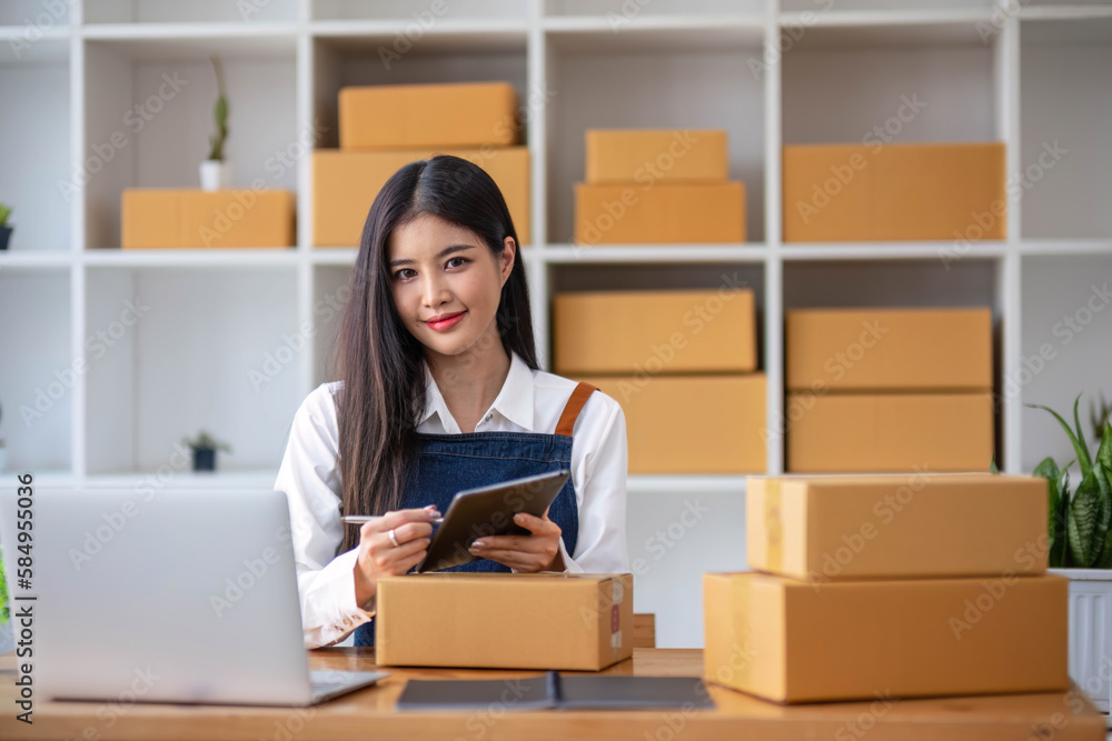 A portrait of a young Asian woman, e-commerce employee sitting in the office full of packages in the background write note of orders and a calculator, for SME business ecommerce and delivery business A portrait of a young Asian woman, e-commerce employee sitting in the office full of packages in the background write note of orders and a calculator, for SME business ecommerce and delivery business