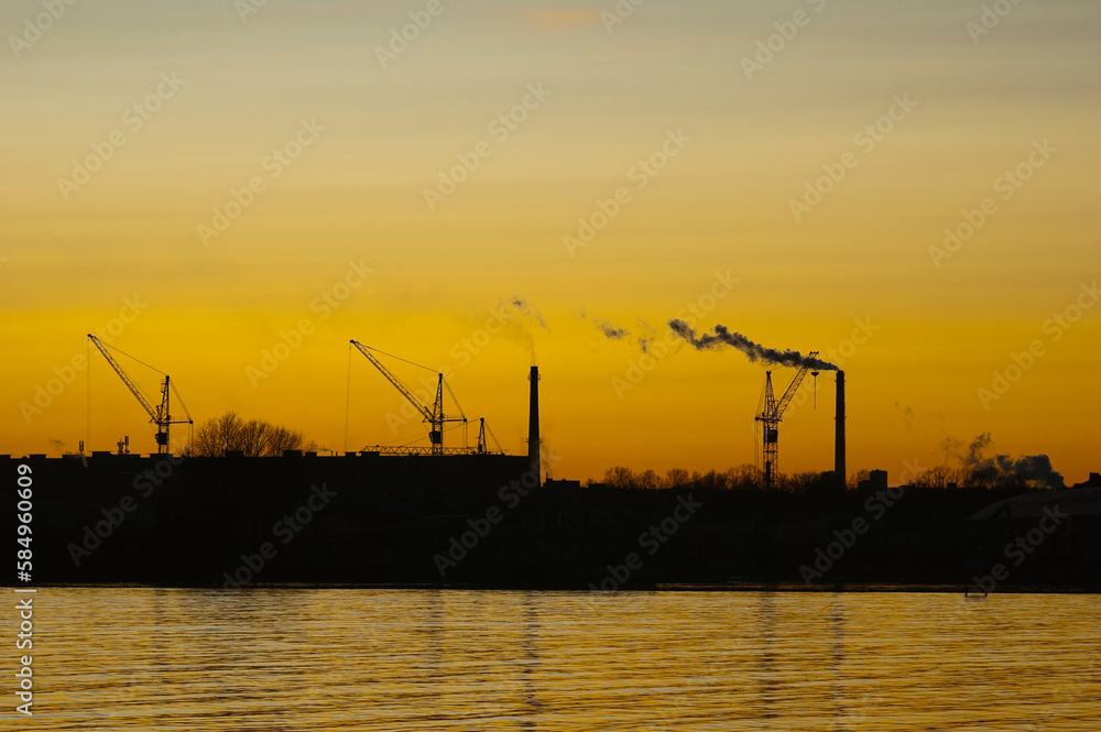 silhouettes of construction cranes and pipes of the plant against the background of the evening sky