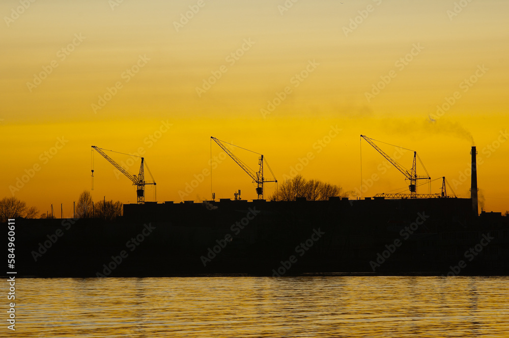 silhouettes of construction cranes and pipes of the plant against the background of the evening sky