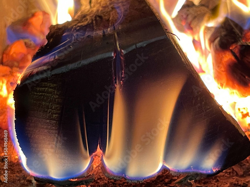 A burning log shows charred wood, glowing embers and rising flames. Blue and orange tones highlight the fire as heat shapes the surface and edges of the wood.