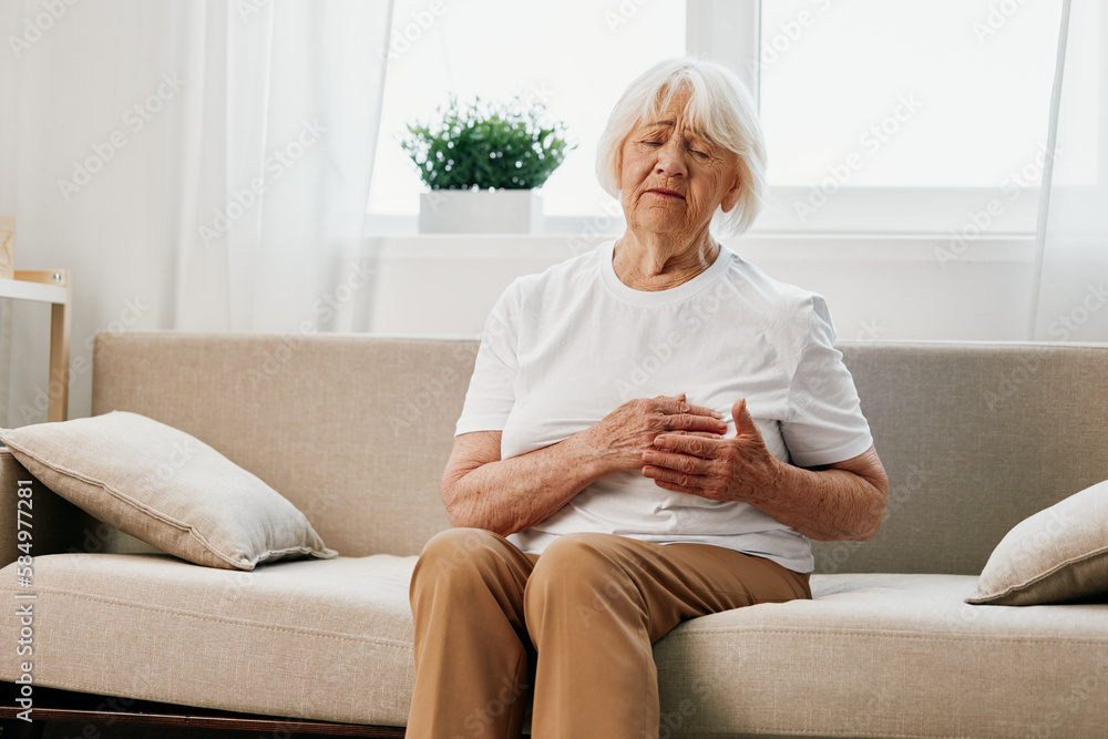 Elderly woman severe pain in the heart sitting on the couch, health problems in old age, poor quality life. Grandmother with gray hair holding on her chest, heart attack and stroke, pressure problem. Elderly woman severe pain in the heart sitting on the couch, health problems in old age, poor quality life. Grandmother with gray hair holding on her chest, heart attack and stroke, pressure problem.