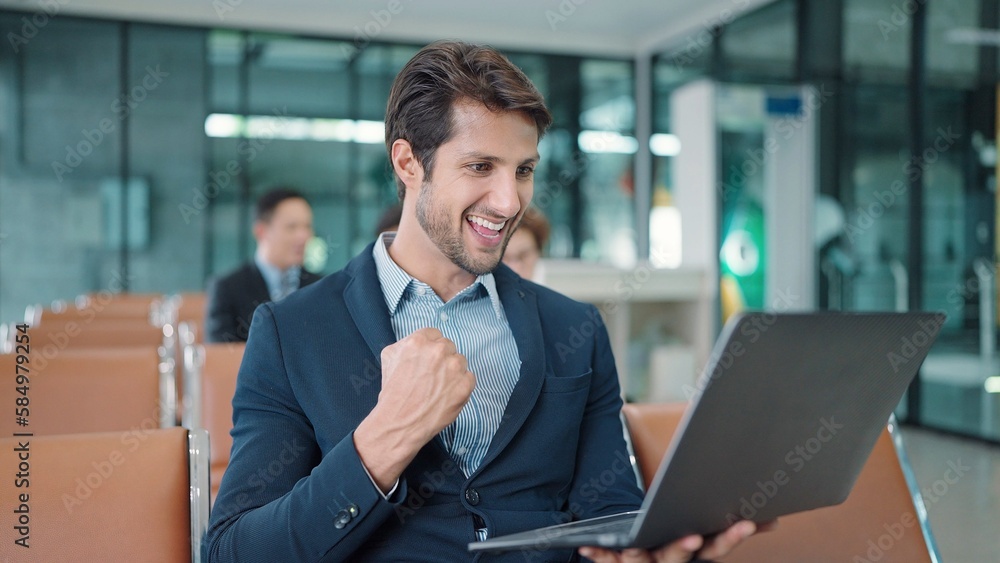 Handsome Indian or latin businessman wearing suit sitting and working on laptop hands up for celebration a good news of work waiting for flight at airport terminal