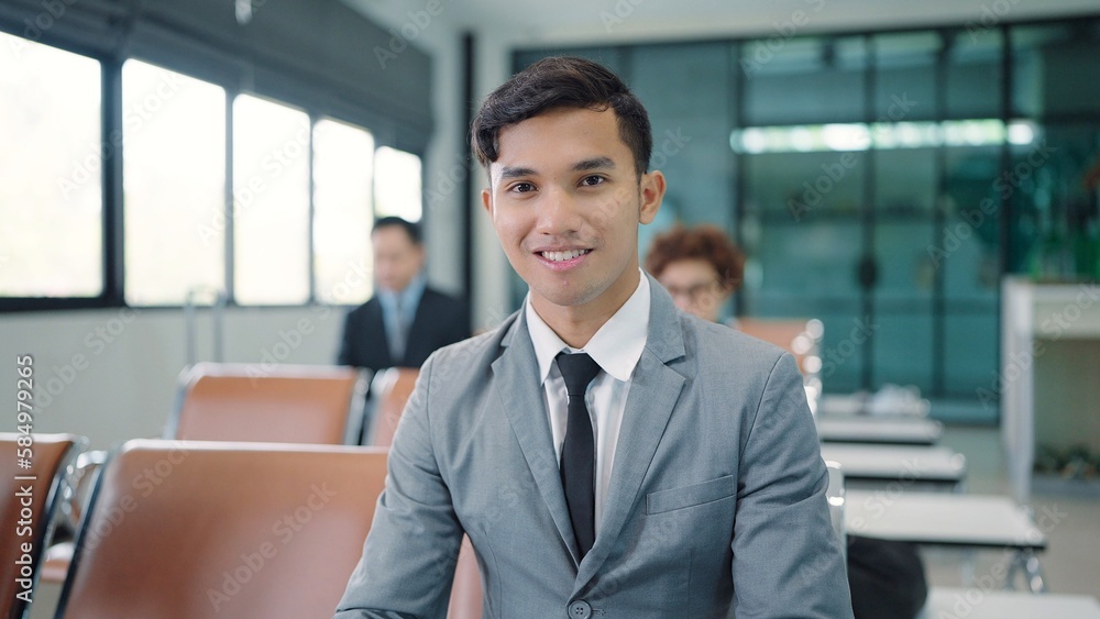 Close up smiling face of young Asian businessman wearing suit sitting at airport terminal looking at camera Close up smiling face of young Asian businessman wearing suit sitting at airport terminal looking at camera