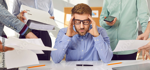 Very tired, stressed and busy man at work. Young man in shirt and glasses sitting at office table, holding his head, and ignoring colleagues giving him lots of business paperwork. Banner background