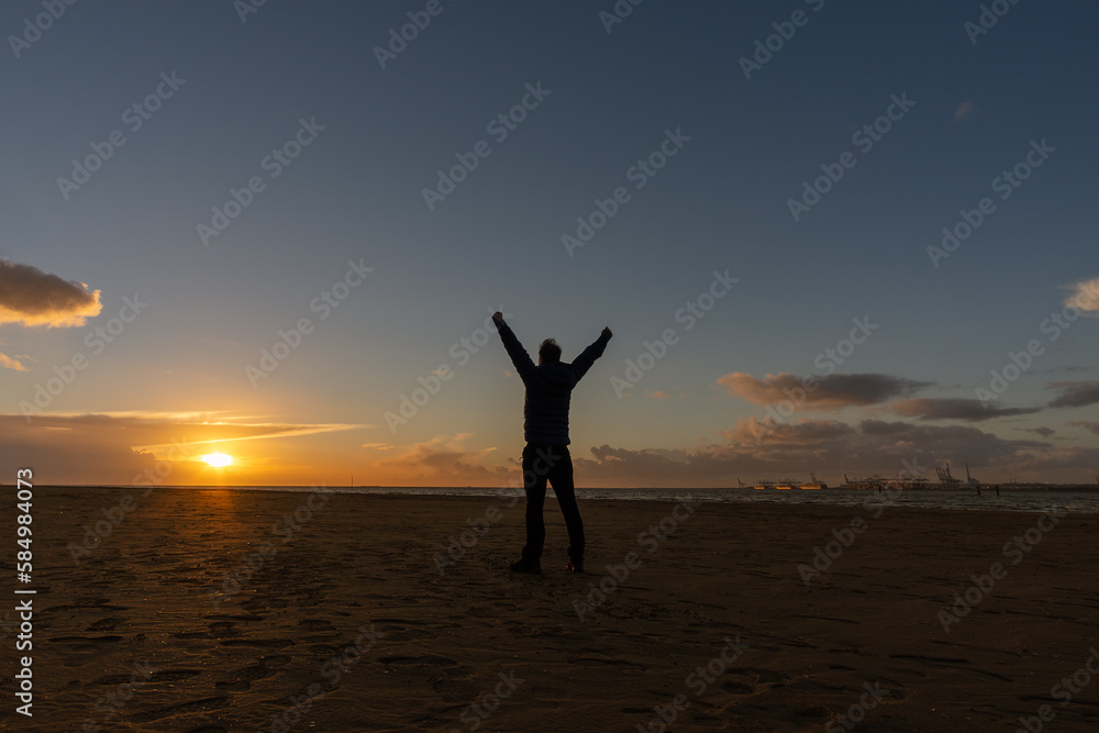 Young man, standing, raising his arms, watching the sunset on beach