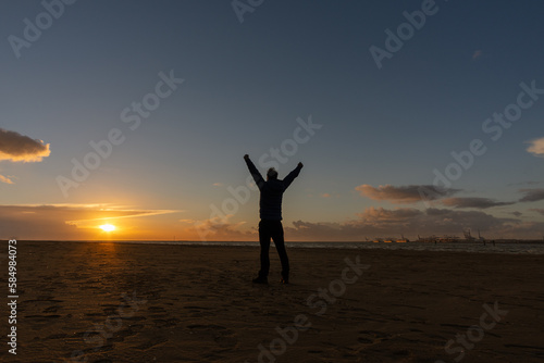 Young man, standing, raising his arms, watching the sunset on beach