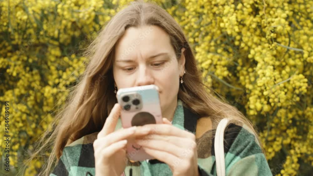A young woman looks at her phone against the background of a blooming yellow mimosa. The wind blows my hair. A young woman looks at her phone against the background of a blooming yellow mimosa. The wind blows my hair.