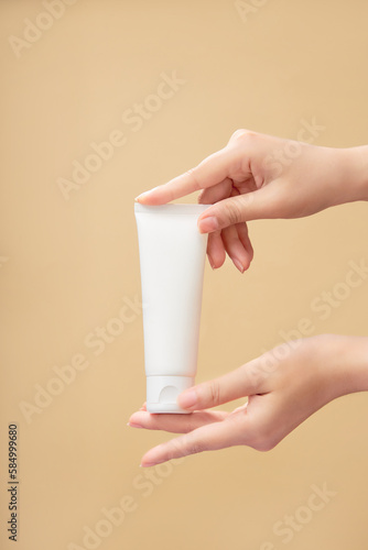 A white empty label tube is held by beautiful woman hands against a beige background. Branding mockup. Natural organic spa cosmetics