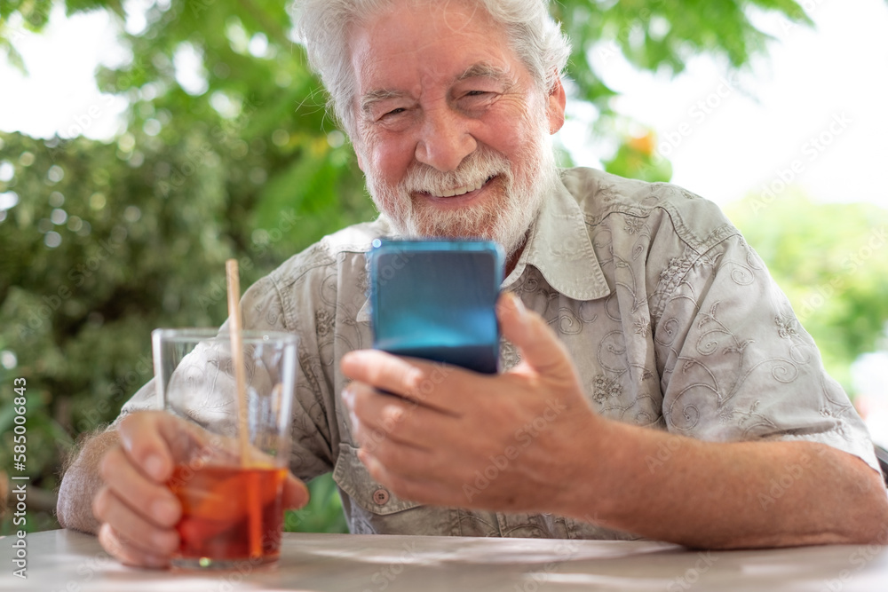 Happy attractive senior man sitting in outdoor in a cafeteria table holding a drinking glass and looking at mobile phone, elderly bearded grandfather relaxing enjoying tech and social Happy attractive senior man sitting in outdoor in a cafeteria table holding a drinking glass and looking at mobile phone, elderly bearded grandfather relaxing enjoying tech and social