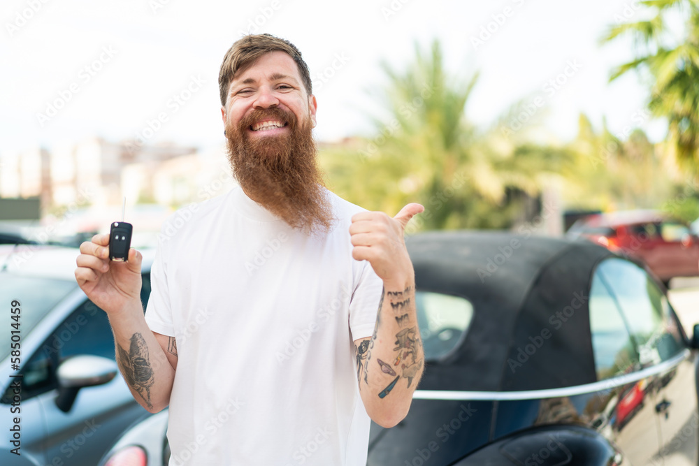 Redhead man with beard holding car keys at outdoors pointing to the side to present a product Redhead man with beard holding car keys at outdoors pointing to the side to present a product