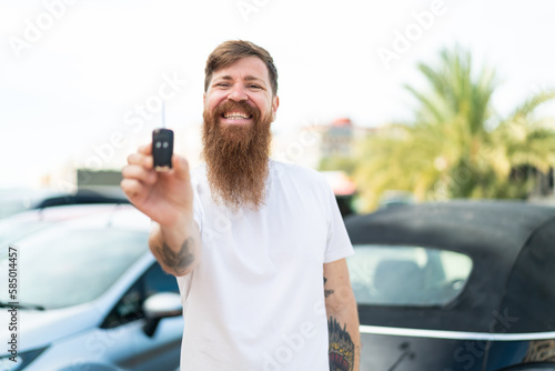 Redhead man with beard holding car keys at outdoors with happy expression