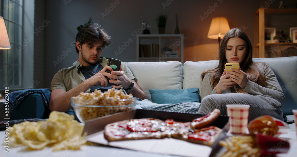 Bored young couple is frustrated after hard day, sitting in front of tv but scrolling through social media on their phones and eating unhealthy food 