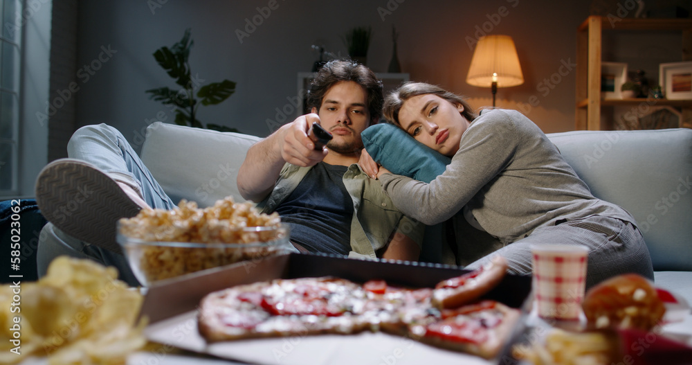 Young caucasian couple extremely frustrated. Bored students are sitting in front of tv, switching channels with remote and eating unhealthy food, tired after hard day Young caucasian couple extremely frustrated. Bored students are sitting in front of tv, switching channels with remote and eating unhealthy food, tired after hard day