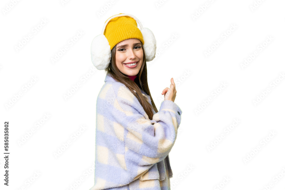 Young woman wearing winter muffs over isolated chroma key background pointing back Young woman wearing winter muffs over isolated chroma key background pointing back