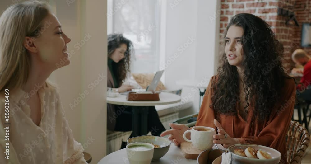 Beautiful young women friends talking and laughing sitting at table in cafe enjoying leisure time together. People and socializing concept. Beautiful young women friends talking and laughing sitting at table in cafe enjoying leisure time together. People and socializing concept.