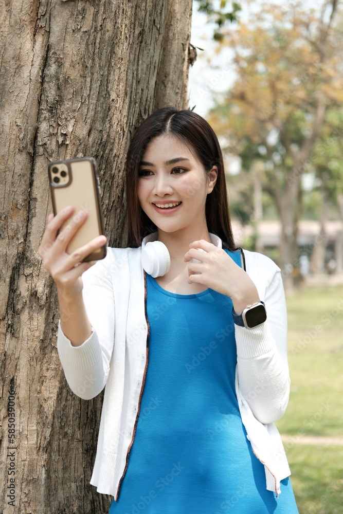 Portrait, Attractive young Asian female in casual clothes using her smartphone while relaxing sitting under the tree in greenery park. Portrait, Attractive young Asian female in casual clothes using her smartphone while relaxing sitting under the tree in greenery park.