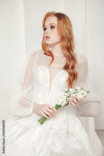 Stylish bride irish lady in white wedding dress with red hair sitting at white wall, looking away. Fashionable cute woman with flowers bouquet. Fashion trendy style beauty concept. Copy ad text space