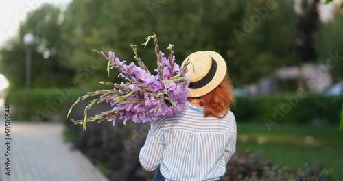 red haired woman in blue shirt and hat walking holding bouquet of purple gladioluses outdoor, summer concept. No face seen. 4k footage photo