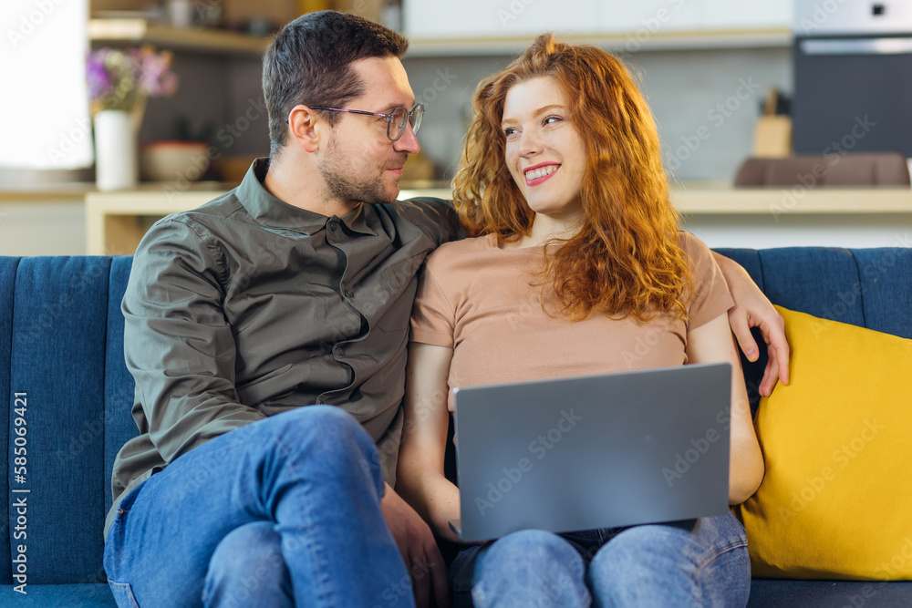 Cheerful cute caucasian couple sitting on sofa and using laptop for internet surf. Living room interior. Cheerful cute caucasian couple sitting on sofa and using laptop for internet surf. Living room interior.