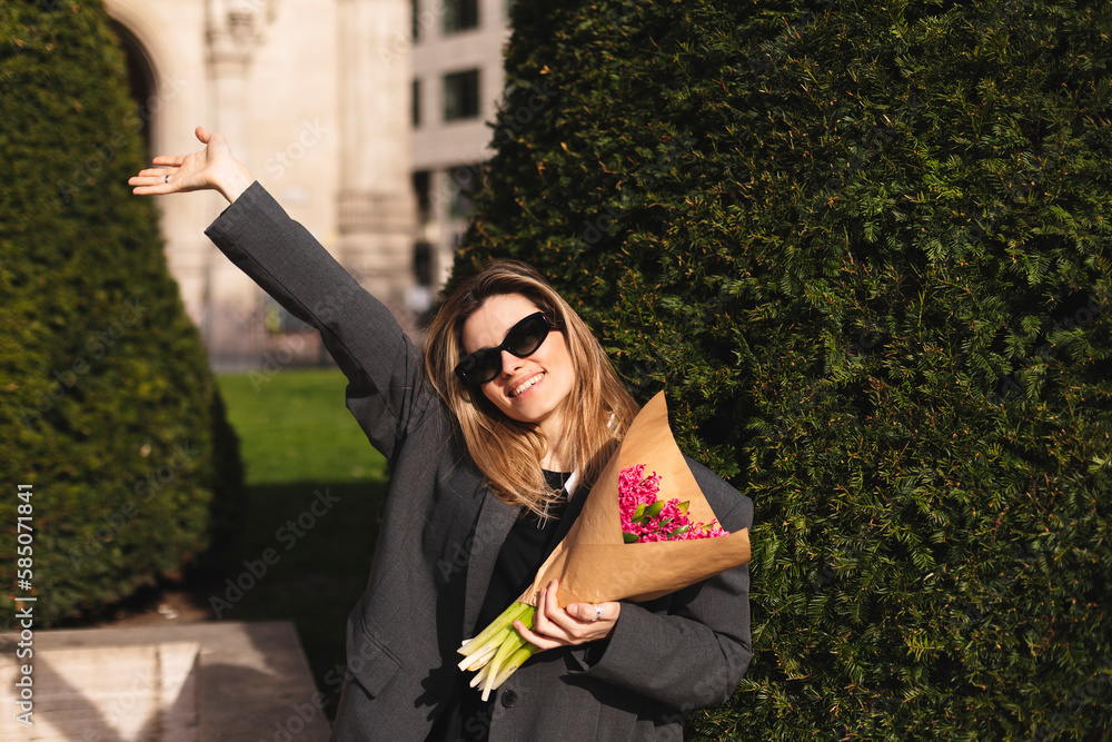 Naklejka premium Happy attractive woman hold bouquet pink flowers walk outdoors, meeting her friends or love, showing high five hey hello hi sign. She is joyful to meet her acquainted by chance. Girl wave her hand.