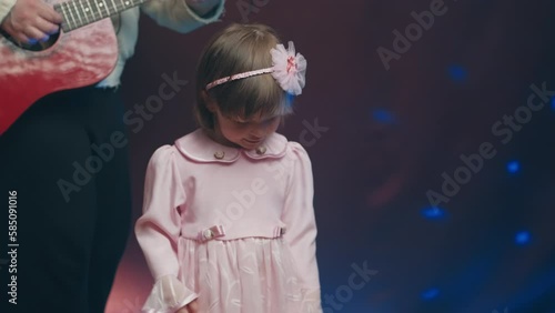 Little girl on stage in pink vintage dress, she dances, her father plays acoustic guitar. Color music is shining. Performance on stage. Early development of children