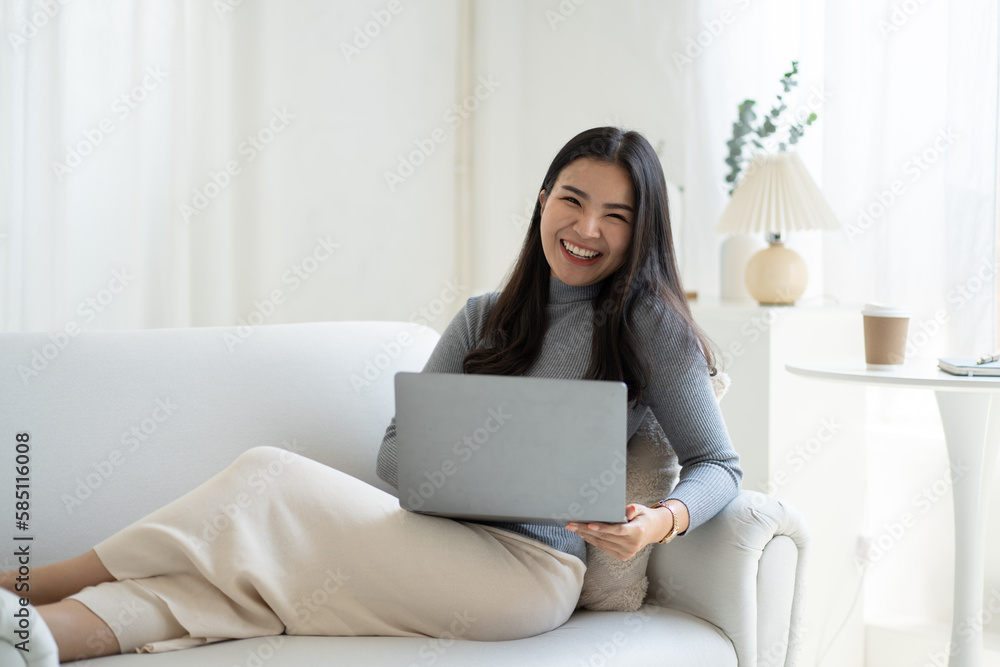 Young beautiful Asian woman using laptop computer while sitting on the sofa at her living room. Young beautiful Asian woman using laptop computer while sitting on the sofa at her living room.