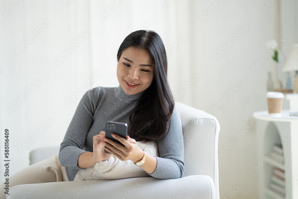 Young attractive Asian female using smartphone while sitting on the sofa in living room. Young attractive Asian female using smartphone while sitting on the sofa in living room.