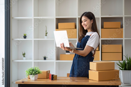 A portrait of a young Asian woman, e-commerce employee sitting in the office full of packages in the background write note of orders and a calculator, for SME business ecommerce and delivery business.