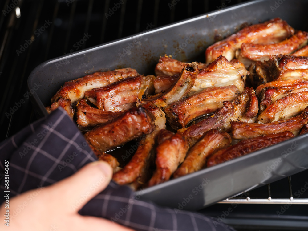 A woman hand taking a black baking tray with baked pork ribs out of an oven.