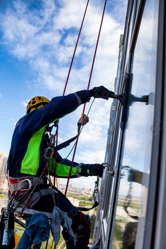 Occupational industrial mountaineering worker washing exterior facade glazing at blue sky backdrop, hanging over building. Rope access laborer climbing on wall of skyscraper. Copy text space for ad