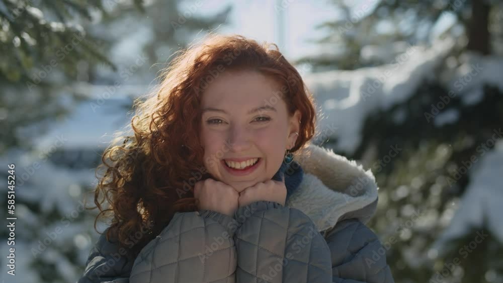A happy red-haired woman smiles at the camera and wraps herself tighter in the collar of her sweater. View of a smiling young woman standing in the middle of snowy trees. A happy red-haired woman smiles at the camera and wraps herself tighter in the collar of her sweater. View of a smiling young woman standing in the middle of snowy trees.